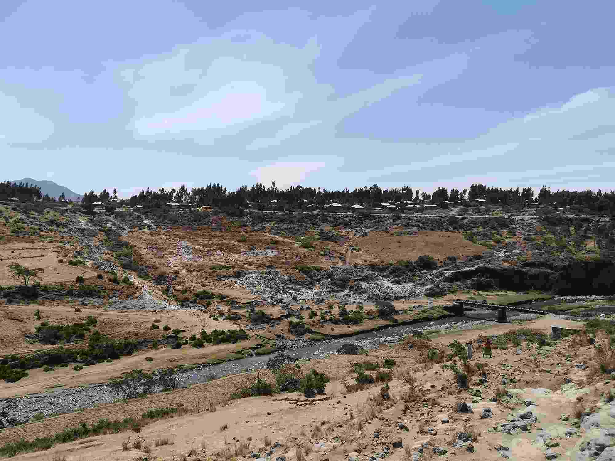 View from a hillside of the Akaki River below, and the dusty brown, hillside rising opposite, scattered with shrubs and trees. 